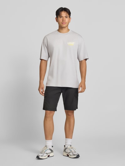 Männermodel trägt Relaxed Fit T-Shirt aus reiner Baumwolle Modell 'Luggage' von Carhartt Work In Progress in Hellgrau, Ganzkörperansicht von vorne. Herren T-Shirt, Label-Detail, Rückenlänge bei Größe M: 78 cm.