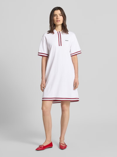Frauenmodel trägt knielanges Polokleid mit Label-Stitching Modell 'Tasa' von JOOP!, Baumwoll-Elasthan-Mix, weiße Grundfarbe, rote Kontraststreifen, gerader Schnitt, Ganzkörperansicht von vorne.
