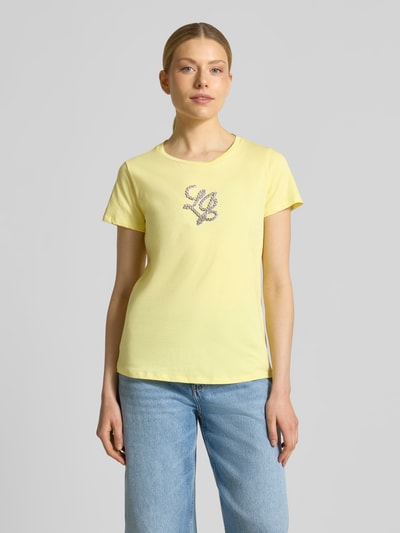 Frauenmodel trägt T-Shirt mit Rundhalsausschnitt und Strasssteinbesatz von LIU JO SPORT aus bawełna, prosty krój, okrągły dekolt, detal z logo z obszyciem kamieniami stras, długość tyłu w rozmiarze S: 60 cm, jasny żółty kolor, gładka tekstura, Ganzkörperansicht von vorne.