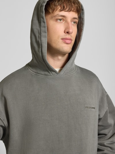 Männermodel trägt Oversized Hoodie aus Baumwoll-Mix von PEGADOR in Grau mit Logo-Stickerei und überschnittenen Schultern, Nahaufnahme Oberkörper.