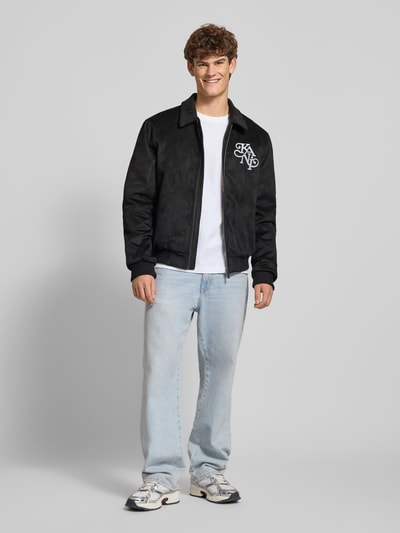 Männermodel trägt Jacke in Leder-Optik mit Label-Stitching von KARL KANI, schwarz, Regular-Fit, Umlegekragen, Reißverschluss, Ganzkörperansicht von vorne. Rückenlänge 68 cm, Schulterbreite 49 cm, Brustweite 59 cm, Ärmellänge 70 cm, Größe M.