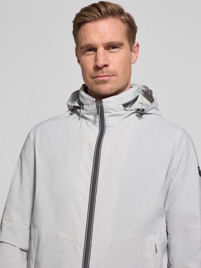 Männermodel trägt Regular Fit Jacke mit opstaande kraag van bugatti in lichtgrijs met Logodetail, capuchon en tweewegritssluiting, Nahaufnahme Oberkörper. Context: Heren jack van bugatti, opstaande kraag, regular fit, Logodetail, capuchon opvouwbaar, tweewegritssluiting, steekzakken, elastische mouwboorden, ruglengte maat 50: 68 cm, schouderbreedte maat 50: 42 cm, borstwijdte maat 50: 50 cm, mouwlengte maat 50: 65 cm.