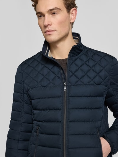 Męski model nosi Slim Fit Steppjacke mit gefüttertem Kragen von Christian Berg Men, granatowy kolor, pikowana tekstura, krój slim fit, ocieplany kołnierz, zasuwane kieszenie, podszewka, elastyczne mankiety, szerokość w rozmiarze M: 52 cm, długość tyłu: 68 cm, szerokość ramion: 47 cm, długość rękawa: 68 cm, widok z przodu, zbliżenie na górną część ciała.
