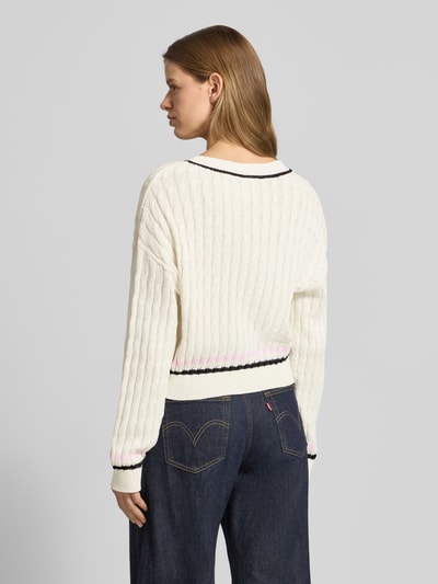 Frauenmodel trägt Strickpullover mit Zopfmuster und V-Ausschnitt Modell 'MAKI' von Noisy May im Baumwoll-Mix, Regular-Fit, Rückenlänge 50 cm, Ärmellänge 55 cm, cremefarben mit schwarzen und rosa Akzenten, grobe Strickstruktur, Ganzkörperansicht Rückansicht.