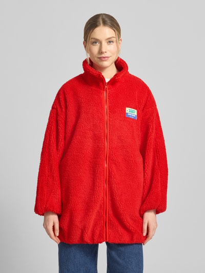 Frauenmodel trägt Oversized Teddyjacke mit Stehkragen Modell 'HOKTOWN' von American Vintage, leuchtend rot, flauschige Textur, Label-Detail, Eingrifftaschen, Reißverschluss, Ganzkörperansicht von vorne. Rückenlänge 89 cm, Schulterbreite 58 cm, Brustweite 67 cm, Ärmellänge 49 cm bei XS/S.