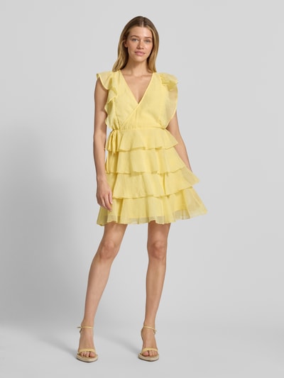 Modelka kobieca nosi tailliertes Cocktailkleid mit Wickeloptik Modell 'SABRINA' od Vero Moda, żółty szyfon, dekolt V, volany, mini, widok całej sylwetki z przodu.