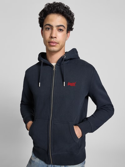 Männermodel trägt Sweatjacke mit Kapuze und Logo-Stitching von Superdry aus Baumwoll-Mix, Regular-Fit, Reißverschluss, Eingrifftaschen, Gerippte-Abschlüsse, Label-Detail, dunkle Farbe, glatte Textur, Ganzkörperansicht von vorne