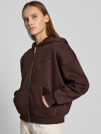 Modelka nosi brązową Relaxed Fit Sweatjacke aus Baumwoll-Mix mit Label-Detail od REVIEW, elastyczne wykończenia, kieszenie kangurki, zamek, bawełniana faktura, widok całej sylwetki z przodu.