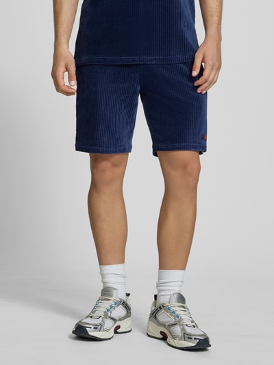Męski model nosi Shorts aus Cord Modell 'TOMATRO' von Ellesse w kolorze granatowym, bawełna z dodatkiem stretchu, krój regular fit, elastyczny pas z tunelowym sznurkiem, szerokość w rozmiarze M: 42 cm, długość wewnętrzna nogawki: 21 cm, faktura sztruksowa, widok całej sylwetki z przodu.