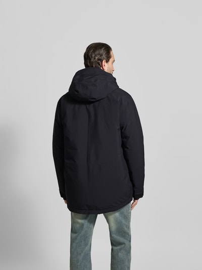 Männermodel trägt Daunenjacke mit Daunen-Feder-Füllung von MM6 Maison Margiela in Schwarz, lockere Passform, Kapuze, Rückenansicht Ganzkörperansicht. Kontext: Herren Daunenjacke, Kinnschutz, Zweiwege-Reißverschluss, Reißverschlusstaschen, Label-Print, verstellbare Ärmelriegel, Tunnelzug im Bund, Rückenlänge 87 cm, Schulterbreite 57 cm, Ärmellänge 38 cm.