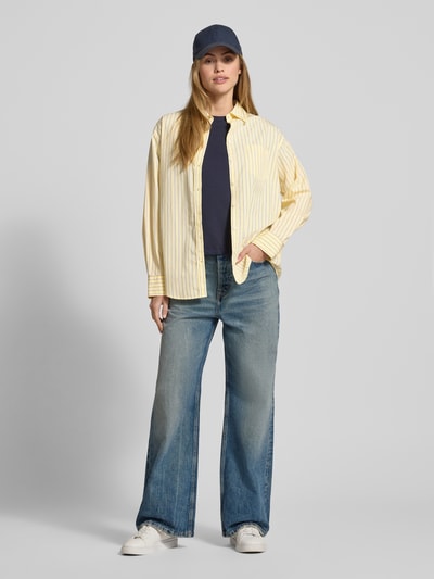 Frauenmodel trägt Relaxed Fit Bluse mit Brusttasche von Gant aus reiner Baumwolle, gelb gestreift, Hemdblusenkragen, Knopfleiste, Ganzkörperansicht von vorne.