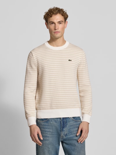 Männermodel trägt Regular Fit Pullover aus Baumwoll-Mix von Lacoste in Creme mit feiner Struktur und weißen Bündchen, Ganzkörperansicht von vorne.