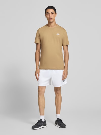 Männermodel trägt Regular Fit T-Shirt aus reiner Baumwolle von Nike in Beige, weiße Shorts, schwarze Schuhe, Ganzkörperansicht von vorne.