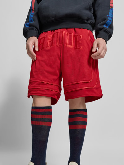 Männermodel trägt Baggy Fit Sweatshorts mit Tunnelzug von RHUDE in Rot, Mesh, Label-Stitching, Eingrifftaschen, elastischer Bund, Nahaufnahme Unterkörper.