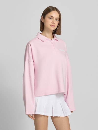 Frauenmodel trägt Sweatshirt mit Label-Print von adidas Sportswear in Hellrosa, lockerer Schnitt, Hemdblusenkragen, Ganzkörperansicht von vorne.
