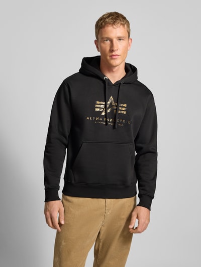 Männermodel trägt Hoodie mit Logo und Känguru-Tasche von Alpha Industries, Baumwoll-Mix, Regular-Fit, schwarze Farbe, Nahaufnahme Oberkörper.