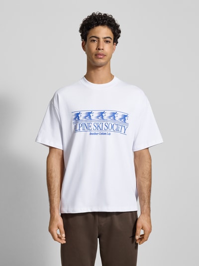 Männermodel trägt T-Shirt mit Label-Print von ANOTHER COTTON LAB, Reine Baumwolle, weiß mit blauem Print, Lockerer-Schnitt, Gerippter Rundhalsausschnitt, Rückenlänge bei Größe M 73 cm, Ganzkörperansicht von vorne.