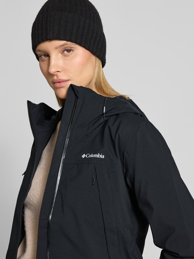 Frauenmodel trägt Regenjacke mit Omni-Tech™ ECO-Technologie von Columbia in Schwarz mit Kapuze, Reißverschlusstaschen und verstellbaren Ärmelriegeln, atmungsaktiv, wasserdicht, Nahaufnahme Oberkörper.