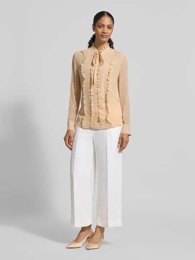 Frauenmodel trägt Regular Fit Bluse aus Seide mit Rüschen von HERZENS in Beige, kombiniert mit weißer Hose, Ganzkörperansicht von vorne.