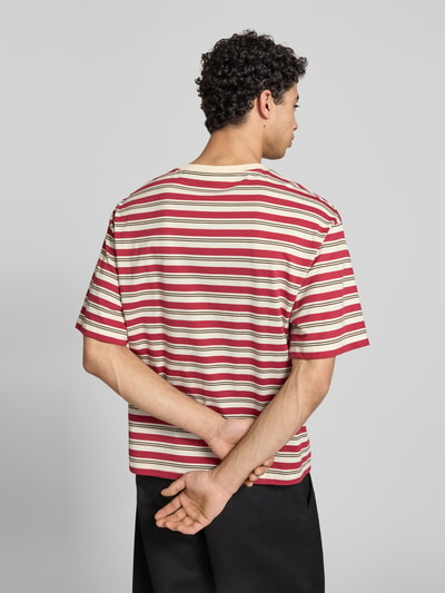 Męski model nosi Relaxed Fit T-Shirt mit Logo-Stitching Modell 'Heritage' von KARL KANI z bawełny, paski czerwone i białe, okrągły dekolt, długość tyłu w rozmiarze M 64 cm, widok całej sylwetki z tyłu.