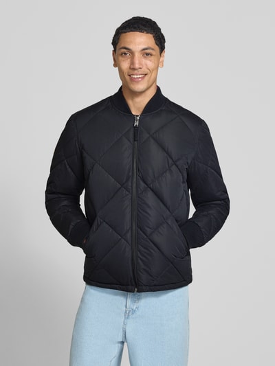 Männermodel trägt Regular Fit Steppjacke mit Stehkragen van Superdry in zwarte Stiksels look met steekzakken, elastische mouwboorden en ritssluiting, Ruglengte maat M 69 cm, schouderbreedte 46 cm, borstwijdte 58 cm, mouwlengte 59 cm, Ganzkörperansicht van voren.