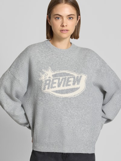 Kobieta modelka nosi sweter z dzianiny 'Strickpullover mit Label-Print' od REVIEW, jasnoszary, luźny krój, okrągły dekolt, nadruk z logo, szeroko opadające ramiona, prążkowany dół, długość tyłu w rozmiarze S: 56 cm, długość rękawa w rozmiarze S: 44 cm, zdjęcie w ujęciu z przodu, zbliżenie na górną część ciała.