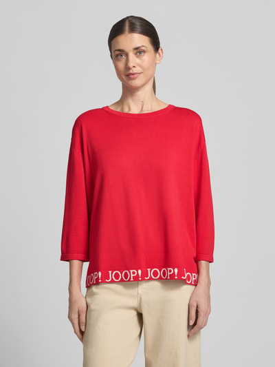 Frauenmodel trägt Strickpullover mit Label-Details und 3/4-Arm Modell 'Kussa' von JOOP!, leuchtend rot, Relaxed Fit, Nahaufnahme Oberkörper.