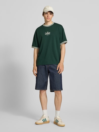 Männermodel trägt Boxy Fit T-Shirt aus reiner Baumwolle Modell 'LES' von Only & Sons in Dunkelgrün, Label Print, Rundhals, unifarben, Kurzarm, ganzkörperansicht von vorne.