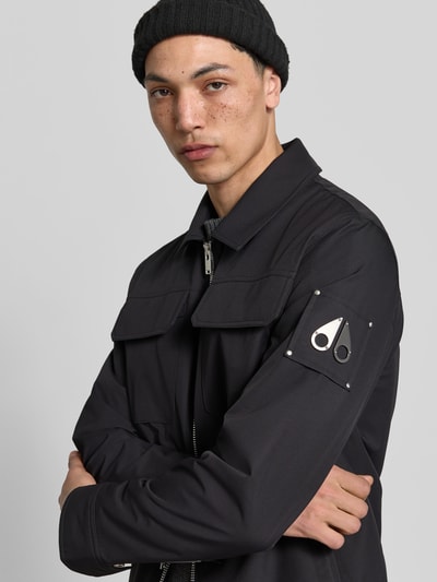 Männermodel trägt Jacke mit Umlegekragen von Moose Knuckles in Schwarz, Brusttaschen, Label-Patch, Zweiwege-Reißverschluss, Nahaufnahme Oberkörper, Gerade Passform, Rückenlänge bei Größe M 67 cm, Schulterbreite 47 cm, Ärmellänge 66 cm.