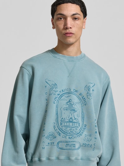 Männermodel trägt Oversized Sweatshirt mit Statement-Print von MAISON KITSUNÉ in Hellblau mit Rundhalsausschnitt und Used-Look, Nahaufnahme Oberkörper.
