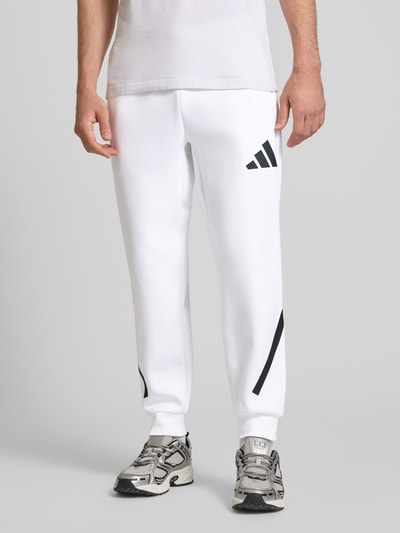 Männermodel trägt Sweatpants mit elastischem Bund von adidas Sportswear in Weiß mit schwarzen Printdetails, reine Baumwolle, Nahaufnahme Unterkörper.