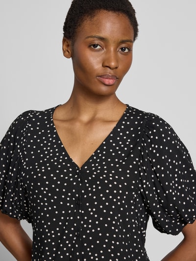 Vrouwenmodel draagt blouse met korte mouwen van zuiver viscose Model 'GILDA' van Soyaconcept, zwarte stof met witte stippen, slim fit, V-hals en knoopsluiting, pofmouwen, ruglengte maat S 33 cm, mouwlengte maat S 27 cm, nahaufnahme bovenlichaam.