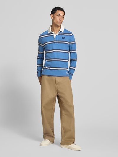 Männermodel trägt Poloshirt aus reiner Baumwolle von Superdry in Blau mit weißen und dunklen Streifen, gerader Schnitt, Polokragen, Ganzkörperansicht von vorne.