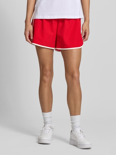 Frauenmodel trägt Track Shorts mit elastischem Bund und Label-Detail von Karo Kauer in Rot mit weißer Kante, Material-Mix mit Stretch-Anteil, Loose-Fit, Bundweite 36 cm, Innenbeinlänge 9 cm, Nahaufnahme Oberkörper.