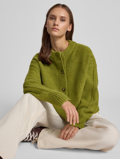 Frauenmodel trägt Boxy Fit Strickjacke Modell 'FLORA' von Topshop in Olivgrün, grob gestrickt, mit Rundhalsausschnitt und Knopfleiste, Damen Strickjacke, Ganzkörperansicht von vorne.