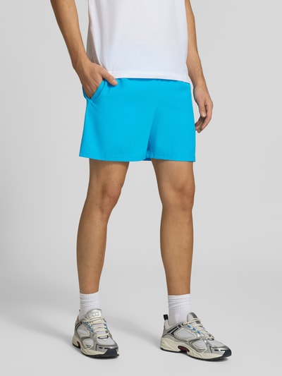 Männermodel trägt Shorts mit elastischem Bund und Logo von New Balance in Blau, glatte Textur, Regular Fit, Nahaufnahme Unterkörper.