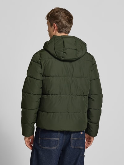 Männermodel trägt Steppjacke mit Kapuze und Reißverschluss von Superdry in Dunkelgrün, wattierte Textur, Ganzkörperansicht Rückansicht, Regular Fit, Stehkragen, verdeckter Reißverschluss, Knopfleiste, Rückenlänge 71 cm, Schulterbreite 50 cm, Brustweite 56 cm, Ärmellänge 83 cm.
