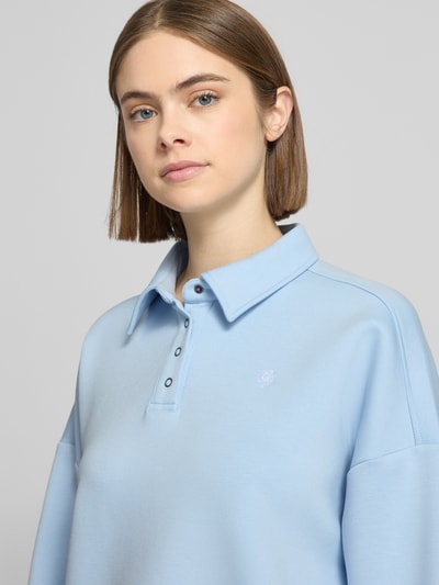 Frauenmodel trägt Cropped Pullover mit Viskose-Anteil von Guess Jeans in Hellblau, Polo-Kragen, Logo-Stitching, Nahaufnahme Oberkörper.