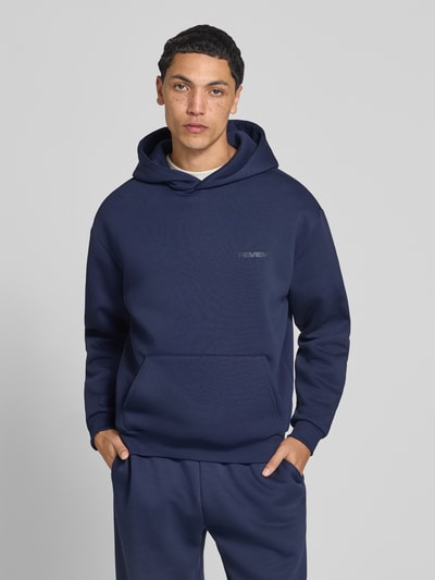 Männermodel trägt Hoodie mit Logo-Print von REVIEW in Dunkelblau, Baumwoll-Mix, Kängurutasche, lockere Passform, Ganzkörperansicht von vorne.