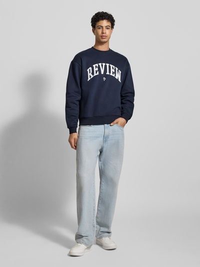 Männermodel trägt dunkelblaues Sweatshirt mit Varsiety Embleme von REVIEW aus Baumwoll-Mix, lockerem Schnitt, Gerippte-Abschlüsse, Rundhalsausschnitt, Ganzkörperansicht von vorne.