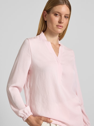 Kobiece modelka nosi Regular Fit Bluse mit V-Ausschnitt von Marc Cain, jasnoróżowa, detal z logo, długość tyłu 65 cm, długość rękawów 59 cm, krój regular fit, dekolt w serek, ganzkörperansicht z przodu.