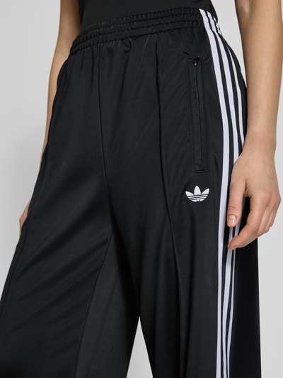 Frauenmodel trägt Sweatpants mit elastischem Bund von adidas Originals in Schwarz mit weißen Streifen, Reißverschlusstaschen, Label-Stitching, Nahaufnahme Oberkörper. Lockerer-Schnitt, Innenbeinlänge bei Größe S: 79 cm.