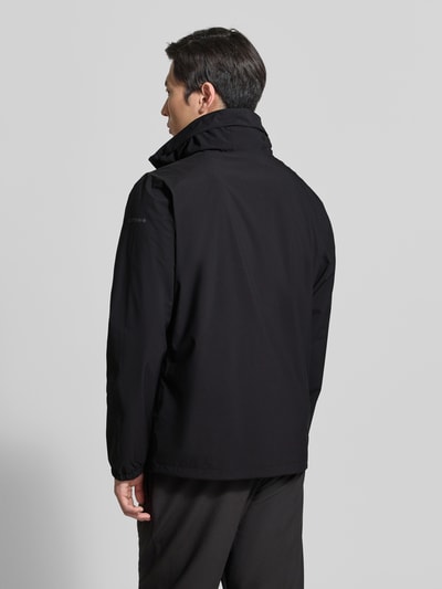 Męski model nosi czarną kurtkę z kapturem model 'BROCTON' od Icepeak, krój regular fit, zamek błyskawiczny, długość tyłu 65 cm, szerokość ramion w rozmiarze 50: 49 cm, szerokość klatki piersiowej w rozmiarze 50: 57 cm, długość rękawów 67 cm, zdjęcie całej sylwetki z tyłu