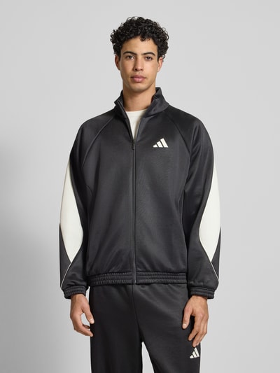 Męski model nosi Kurtka treningowa z logo i stójką od adidas Sportswear, mieszanka materiałów z bawełną, krój regular fit, czarna z białymi wstawkami, zamek błyskawiczny, długość tyłu 68 cm, długość rękawa 67 cm, widok całej sylwetki z przodu.