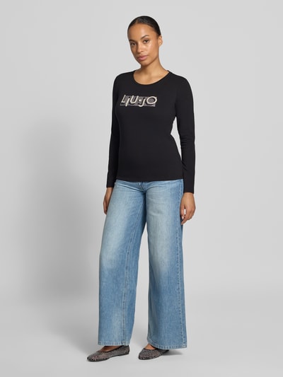 Frauenmodel trägt Langarmshirt aus Baumwoll-Mix von Liu Jo mit Label-Print und Ziersteinen, schwarze Farbe, Ganzkörperansicht von vorne.