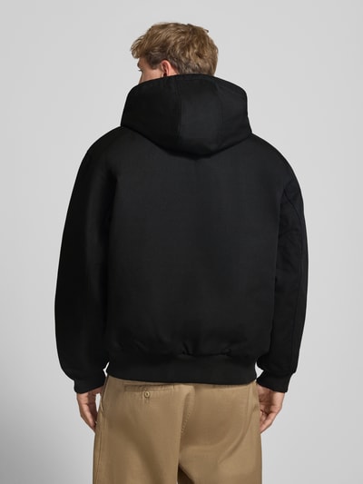 Männermodel trägt Jacke mit Label-Patch und Känguru-Tasche von Carhartt Work In Progress in Schwarz, glatte Baumwollstruktur, Ganzkörperansicht Rückansicht.