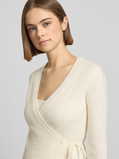 Kobiece modelka nosi Kardigan in Wickel-Optik od Gina Tricot, mieszanka materiałów z domieszką wełny z alpaki, krój slim fit, dekolt w serek, beżowy kolor, tekstura miękka, kopertowy styl, zdjęcie z przodu, ujęcie zbliżeniowe na górną część ciała.