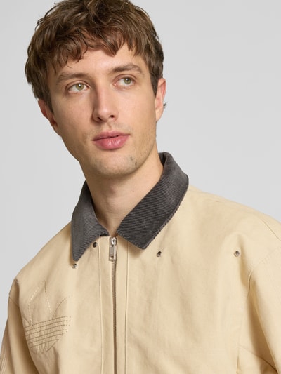 Männermodel trägt Regular Fit Jeansjacke aus reiner Baumwolle von adidas Originals in Beige mit Umlegekragen, Nahaufnahme Oberkörper.