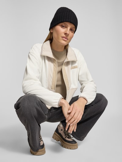 Modelka nosi Cropped Fleecejacke mit Label-Patch Modell 'Backbowl™' od COLUMBIA, prosta forma, stójka, długi zamek, białe i beżowe kolory, elastyczne mankiety, kieszenie, szerokość w rozmiarze S: 57 cm, długość tyłu: 52 cm, szerokość ramion: 55 cm, długość rękawa: 56 cm, widok całej sylwetki z przodu