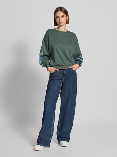 Frauenmodel trägt Relaxed Fit Sweatshirt aus reiner Baumwoll-Mix Modell 'BROOKE' von ONLY, dezentes Grün mit floralen Mustern am Ärmel, kombiniert mit blauer Jeans, Damen Sweatshirt von ONLY, Ganzkörperansicht von vorne.